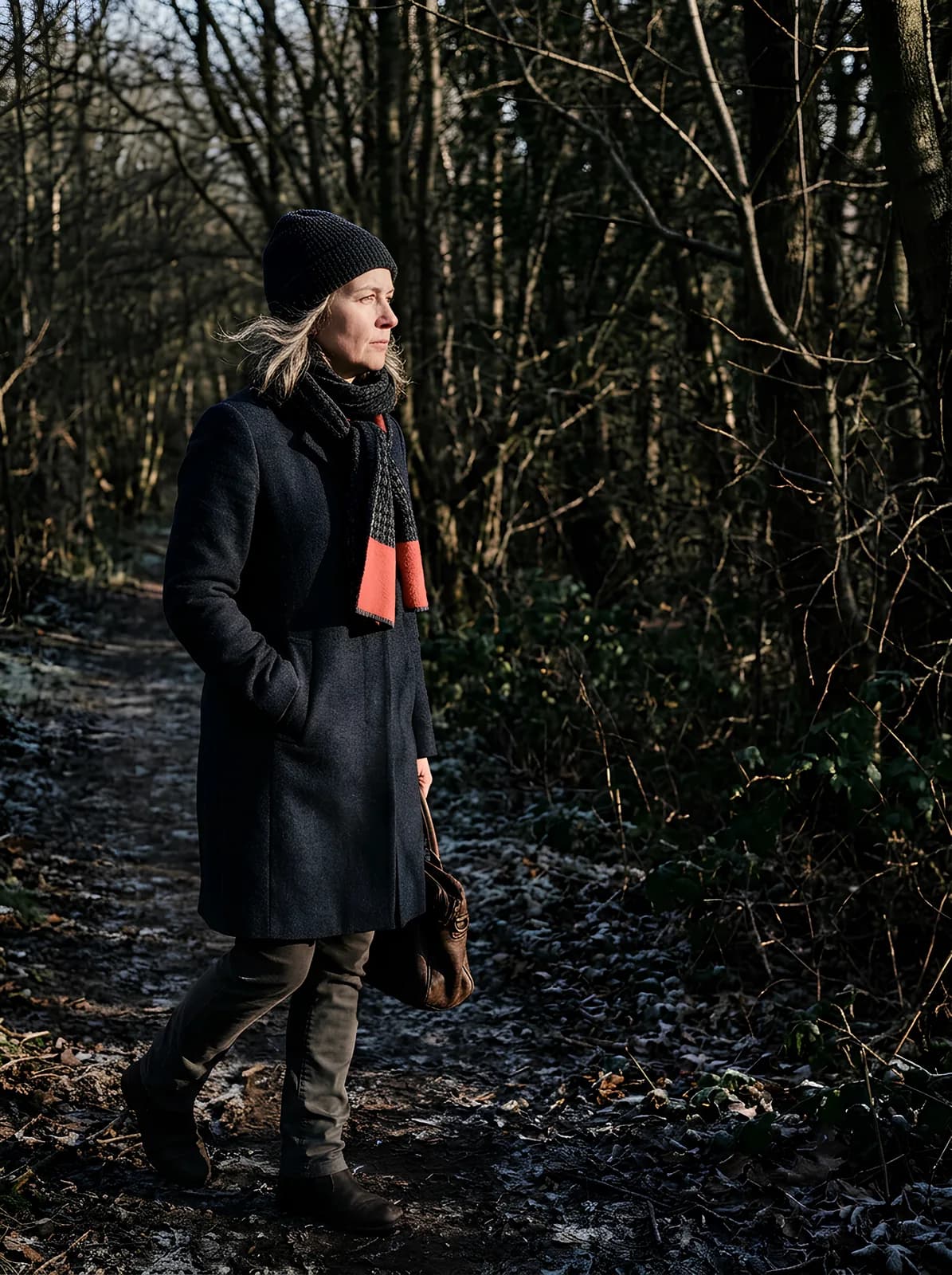A woman in her fifties walking a frost-covered woodland path in low winter sun, caught mid-step with her head turned to look at something off to her right. She wears a dark charcoal wool coat, a ribbed grey beanie and a grey scarf with a single coral panel, with a brown leather bag at her side and one hand in her coat pocket. Bare winter branches fill the dark background, and hard low light rakes across her face from the left.